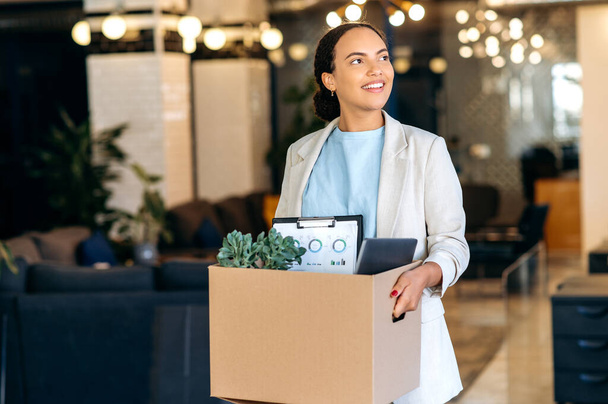 Un nuevo trabajo. Emocionada mujer de raza mixta motivada con éxito, entra en una nueva oficina de trabajo, sostiene una caja de cartón con atributos de trabajo en sus manos, inspecciona el espacio de trabajo con asombro, sonriendo felizmente - Foto, imagen