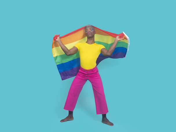 Hombre afroamericano descalzo de cuerpo completo en ropa colorida con cabeza cubierta de maquillaje con bandera LGBT contra fondo turquesa - Foto, Imagen