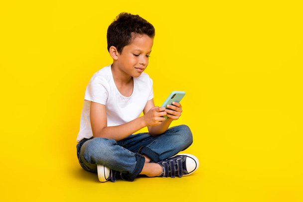 Plná velikost fotografie malého chlapce sedět vzhled telefon nošení trička džíny boty izolované na žluté barvy pozadí. - Fotografie, Obrázek