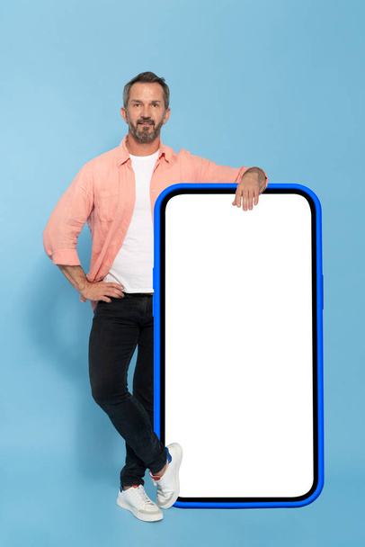 Middle aged fit man leaned on huge, big smartphone with white screen in blue case happy smiling on camera wearing peach shirt and black jeans isolated on blue background. Mobile app advertisement.  - Photo, Image