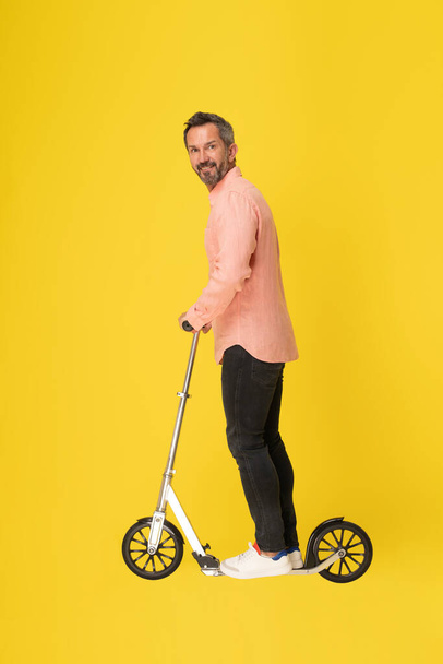 Middle aged grey haired man ride scooter standing sideways looking at camera wearing peach shirt and black jeans isolated on yellow background. Fit masculine handsome mature businessman on the go. - Фото, зображення