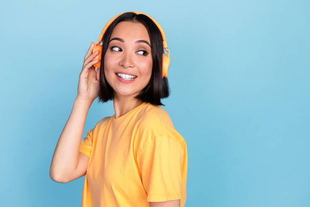 Photo of pretty positive girl hand touch headphones toothy smile look empty space isolated on blue color background. - 写真・画像
