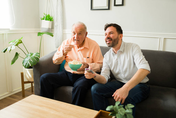 Jovem alegre sorrindo enquanto assiste a um filme de TV com seu pai idoso no sofá enquanto come pipocas - Foto, Imagem