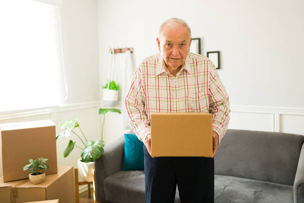 Homem idoso caucasiano sênior fazendo contato visual enquanto carrega uma caixa e se muda para uma nova casa ou apartamento - Foto, Imagem