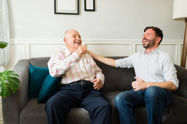 Cheerful young man having fun and laughing with his happy elderly father while chatting together in the living room - Photo, Image