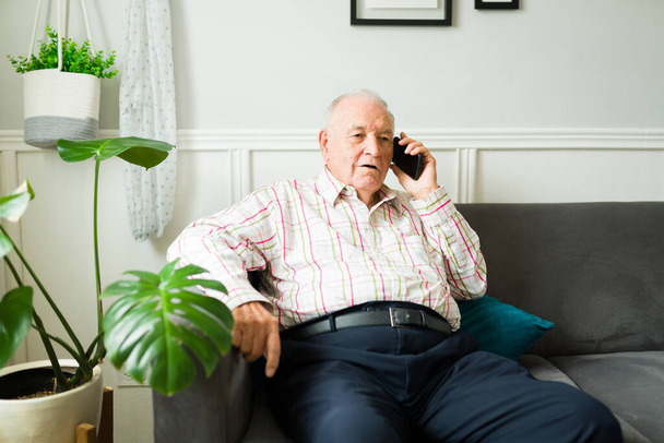 Kaukasischer alter Mann sieht entspannt auf dem Sofa sitzend und ausgeruht aus, während er mit Familie oder Freunden telefoniert - Foto, Bild