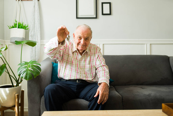 Веселый пожилой человек показывает свои новые ключи от квартиры после переезда домой и выглядит очень счастливым - Фото, изображение