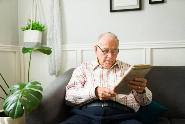 Kluger älterer Herr liest Zeitung und Nachrichten, während er es sich auf dem Wohnzimmersofa gemütlich macht  - Foto, Bild