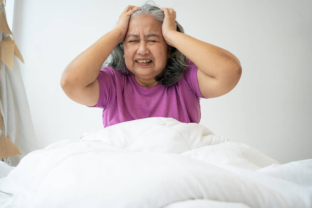 Unhealthy elderly senior Asian woman sitting on bed suffering from headache and migraine so pain and illness, Stress senior woman sit on bed at home alone, tried, Unhappy Elderly despond concept - Фото, изображение
