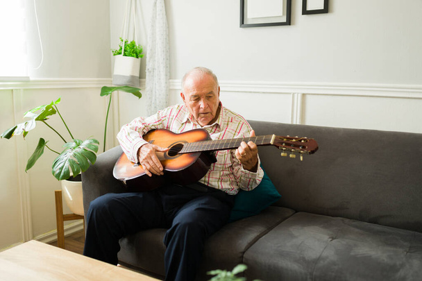 Talented old senior man playing the guitar and sining a song while enjoying his music sitting on the sofa - Foto, afbeelding