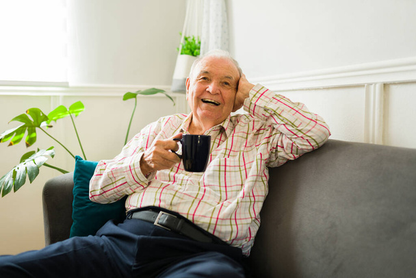 Ενθουσιασμένος ηλικιωμένος καυκάσιος άντρας να γελάει ενώ δείχνει χαλαρός πίνοντας ένα φλιτζάνι καφέ και ξεκουραζόμενος στον καναπέ στο σπίτι - Φωτογραφία, εικόνα