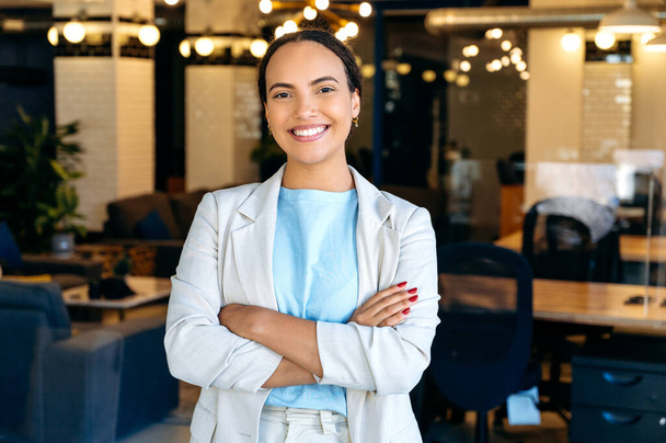 Portrait of a confident successful modern mixed race business lady, top manager of a company, standing in her modern creative office, wearing formal wear, arms crossed, looking at the camera, smiling - Photo, Image