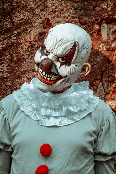 gros plan d'un clown maléfique effrayant, vêtu d'un costume gris avec une ruche blanche et des pompons rouges, fixant l'observateur avec un sourire effrayant, devant le mur extérieur rouillé d'une maison abandonnée au crépuscule - Photo, image