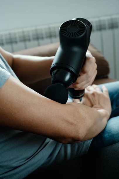 Ένας νεαρός Καυκάσιος χρησιμοποιεί ένα όπλο μασάζ για να κάνει μασάζ στους μυς του χεριού του δίπλα στον αγκώνα του, καθισμένος σε έναν καναπέ. - Φωτογραφία, εικόνα