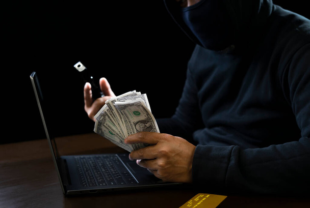 concepto de delito cibernético, ladrón en sudadera con capucha negro que sostiene el dinero y el arma en las manos y el ordenador portátil con tarjeta de crédito puesta en la mesa sobre fondo oscuro - Foto, imagen