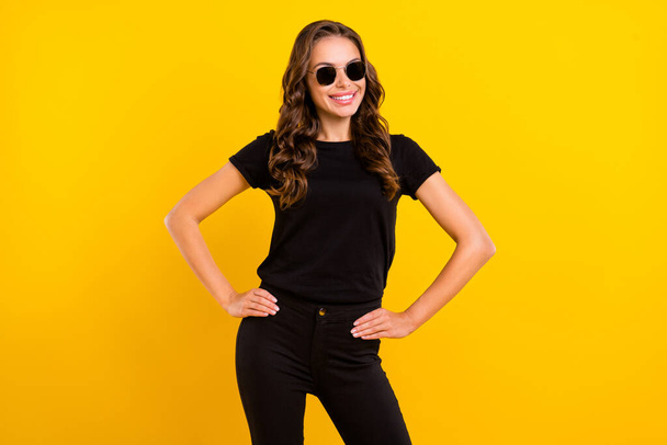 Photo of pretty adorable young lady dressed black clothes dark eyewear arms waist smiling isolated yellow color background. - Fotó, kép