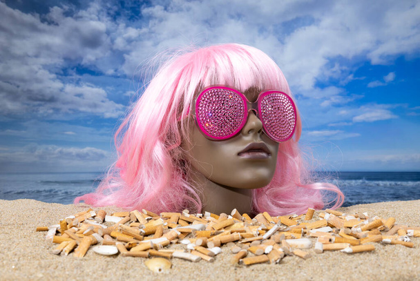 Una cabeza de maniquí femenino de plástico atascado en la arena con la playa llena de colillas de cigarrillos sucios  - Foto, Imagen