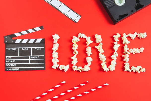 Film clapperboard, film strip, videokazety, brčka a slovo láska z popcornu leží na červeném pozadí, horní pohled - velký plán. Kino láska a Valentýna koncept. - Fotografie, Obrázek