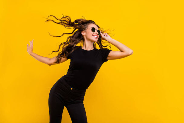 Photo of cool adorable young lady dressed black clothes dark eyewear wind blowing dancing smiling isolated yellow color background. - Fotografie, Obrázek