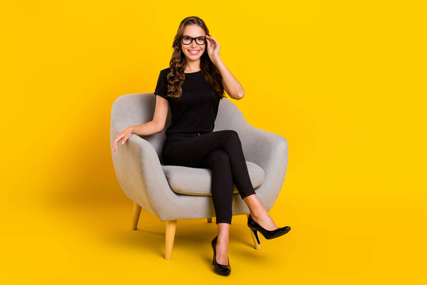 Photo of adorable thoughtful young lady dressed black clothes eyewear sitting grey chair smiling isolated yellow color background. - Foto, Bild