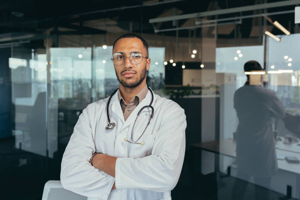 Portrait of serious and successful doctor, man in medical gown looking thoughtfully at camera, working inside modern office building. - Фото, изображение