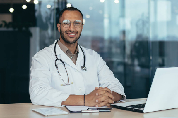 African american doctor portrait, man working inside modern clinic office at table using laptop, doctor in medical coat and stethoscope smiling and looking at camera. - 写真・画像