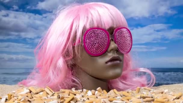 Une tête de mannequin femelle en plastique coincée dans le sable avec la plage avec des mégots de cigarette empilant - Séquence, vidéo