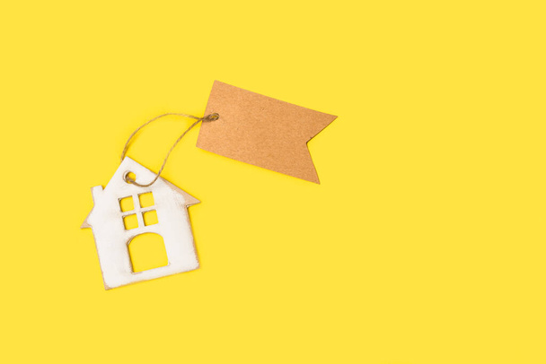 Картонный дом с коричневой пустой бумажной этикеткой на желтом фоне с копировальным пространством - Фото, изображение
