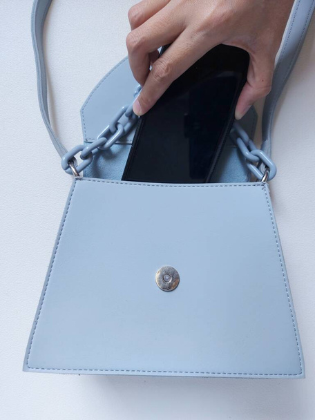 Női parittya táska, kék színű lánccal. mobiltelefonnal és női kezekkel kiegészítve. Elszigetelt háttér fehér - Fotó, kép