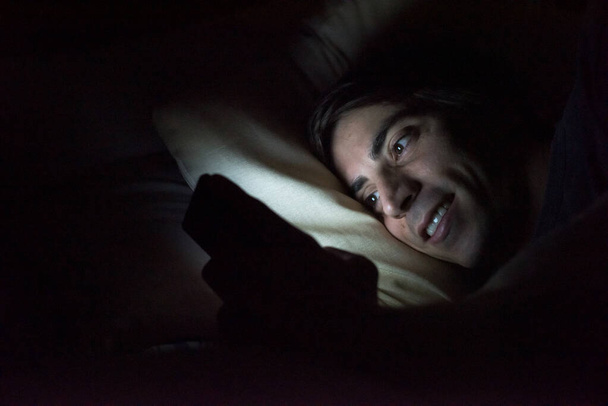 Άγρυπνος νεαρός στο κρεβάτι στο σπίτι χρησιμοποιώντας smartphone τη νύχτα ελέγχοντας τα μέσα κοινωνικής δικτύωσης - Φωτογραφία, εικόνα