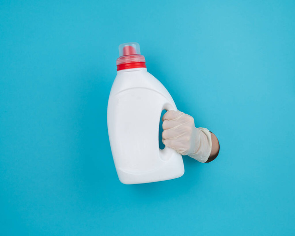 A woman is holding a white bottle of detergent. Womans hand sticking out of blue paper background - Foto, Imagem