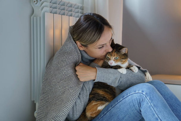 Polish woman with dark circles under eyes hugs and kisses favourite cat. Young female owner looks tired sitting together with pet on floor. Lady covered with warm blanket enjoys spending time with cat - Foto, Imagen