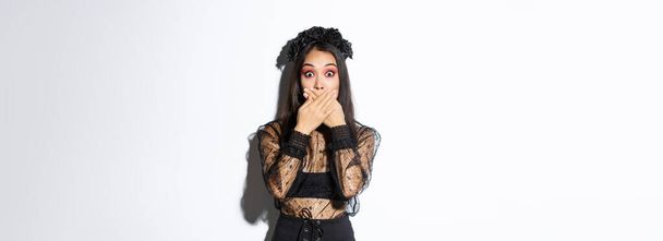 Shocked woman in witch outfit gasping and cover mouth speechless, standing over white background, looking at something surprising, wearing black lace dress and wreath, white background. - Photo, image