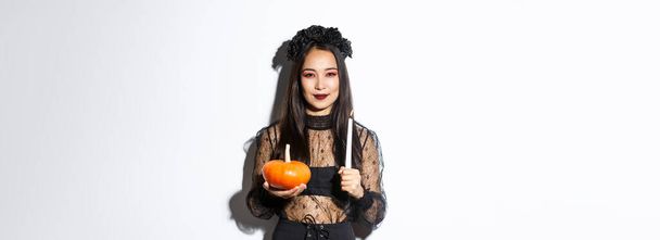 Gotik elbiseli sinsi Asyalı cadının resmi, balkabağıyla mum tutuyor ve kameraya kurnaz bir şekilde bakıyor, beyaz arka planda duruyor.. - Fotoğraf, Görsel
