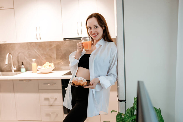 Mujer sonriente alegre bebiendo jugo y comiendo croissant en la cocina casera. concepto de estilo de vida saludable - Foto, imagen
