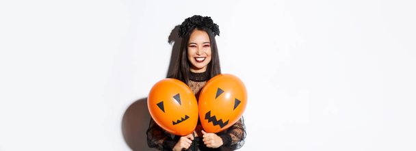 Immagine di ragazza asiatica in costume da strega malvagia con due palloncini arancioni con facce spaventose, che celebrano Halloween, in piedi su sfondo bianco. - Foto, immagini