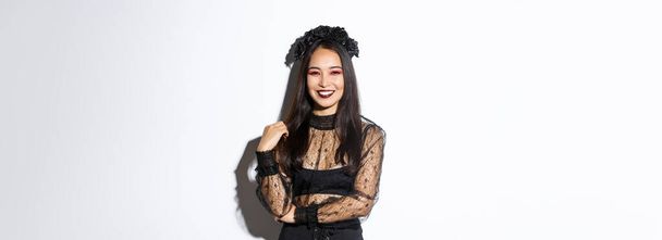 Beautiful young happy woman enjoying halloween party, smiling and looking cheerful while wearing her evil witch costume for trick or treating, standing over white background. - Foto, Imagem