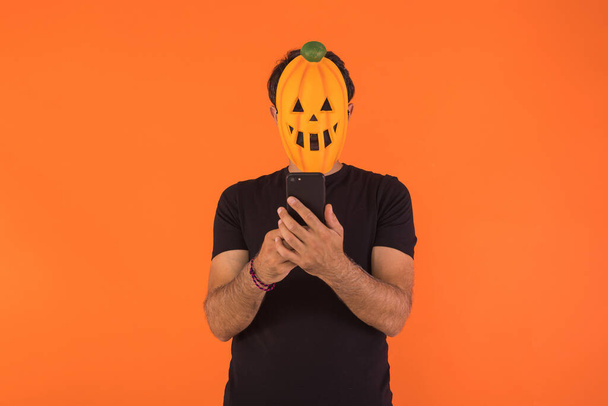 Cadılar Bayramı 'nı kutlayan balkabağı maskeli bir adam turuncu arka planda cep telefonuna bakıyor. Kutlama kavramı, Ruhlar Günü ve Azizler Günü.. - Fotoğraf, Görsel