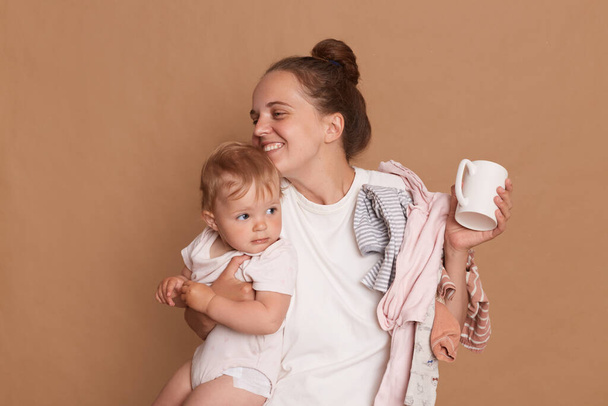 Innenaufnahme einer liebenden jungen Mutter mit Dutt-Frisur, die ihre kleine Tochter umarmt, Tasse in den Händen hält, Glück ausdrückt, isoliert vor braunem Hintergrund posiert. - Foto, Bild