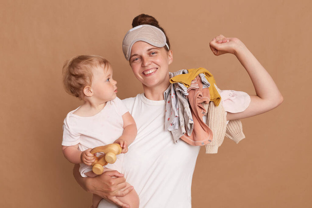 Horizontal shot of smiling woman with dark hair wearing white t shirt and blindfold standing with her baby daughter, stretching her arm, waking up in good mood, posing isolated over brown background. - Foto, imagen
