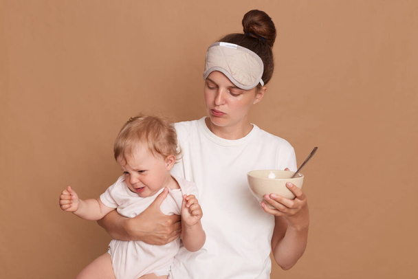 Horizontal shot of woman wearing white t shirt and sleeping mask, mother holding crying baby daughter and plate with kid's food in hands, posing isolated over brown background. - Foto, afbeelding