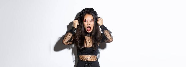 Angry asian woman impersonate evil witch on halloween party, looking mad and furious, screaming to scare people, standing over white background in gothic dress. - Photo, Image