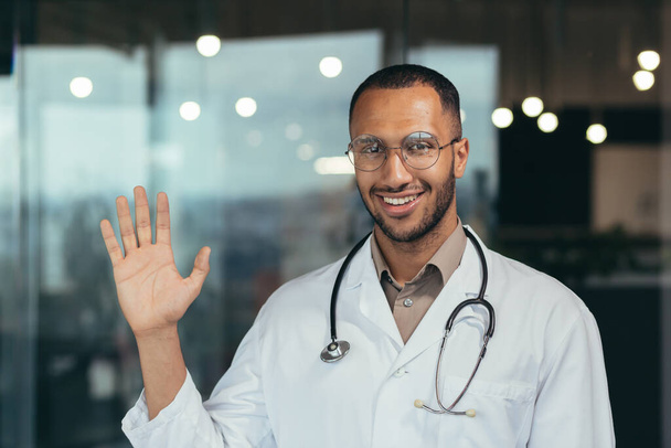 Young african american doctor man wearing glasses wearing medical coat saying hello happy and smiling friendly welcoming gesture doctor working inside clinic. - Zdjęcie, obraz