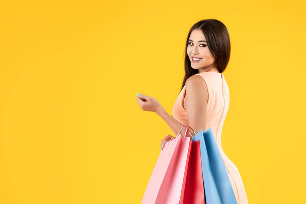 Pretty asian woman holding shopping bags on a yellow background, looking at camera, half length. Summer sales concepts. - Foto, imagen