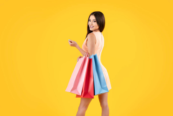 Pretty asian woman holding shopping bags on a yellow background, looking at camera, copy space. Summer sales concepts. - Fotoğraf, Görsel