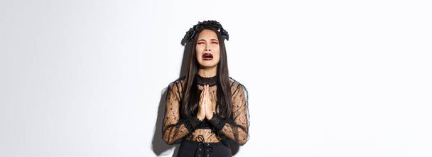 Nieszczęsna młoda azjatycka kobieta w tarapatach błagając Boga, płacząc i błagając o pomoc, nosząc halloween gotycką sukienkę i wieniec, błagając na białym tle. - Zdjęcie, obraz