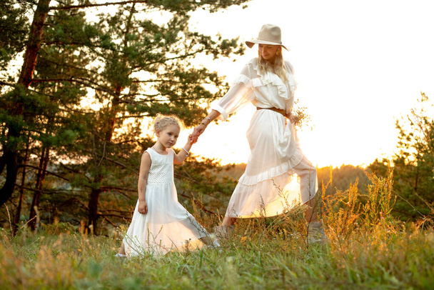 Όμορφη γυναίκα, μητέρα με καπέλο και μικρή ξανθιά κόρη με ανοιχτόχρωμα λευκά φορέματα που περπατούν μαζί στο φυσικό δάσος με θέα το ηλιοβασίλεμα. Οικογενειακή σχέση δεσίματος και αγάπης. Διασκεδάζοντας και περνώντας χρόνο - Φωτογραφία, εικόνα