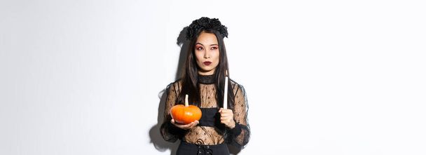 Image of asian woman in wicked witch costume, looking left serious, holding lit candle and pumpkin, celebrating halloween. - Foto, Imagen