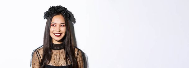 Close-up van mooie elegante vrouw in heks kostuum en gotische make-up, glimlachen blij terwijl het kijken naar de linker bovenhoek, vieren Halloween. - Foto, afbeelding