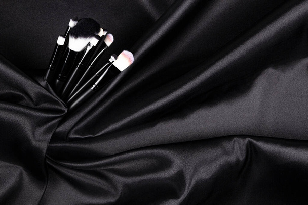 Make-up cosmetic brushes on black wavy satin silk background. Beauty, skin care and fashion composition. Top view. - Foto, Bild